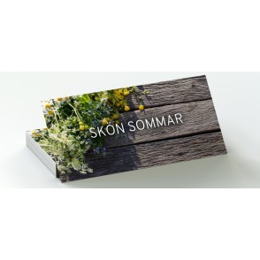 En bild på att rektangulärt gåvokort med texten "Skön Sommar". Texten på gåvokortet ligger mot en härlig bakgrund som visar en träbänk med sommarblommor på.